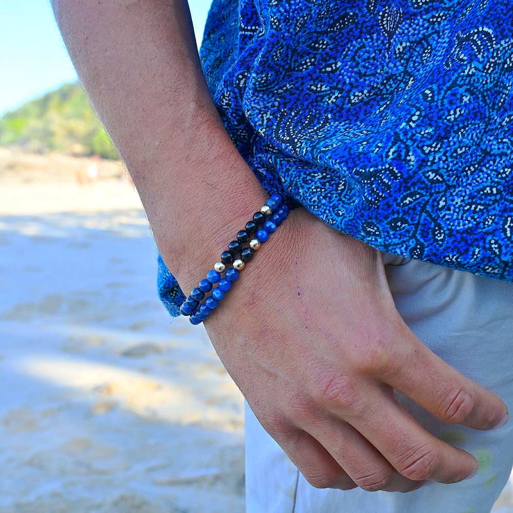 Close-up of a man wearing two stacked Manifestation beaded bracelets (Mastering My Own Reality and Seek & You Shall Discover) with deep blue and black beads, acting as functional tools for intentional creation and unlocking personal power.