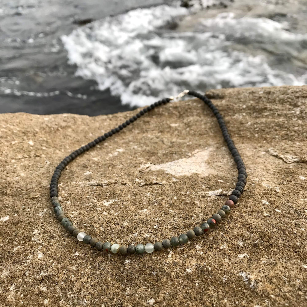 Conviction men's necklace: dark bloodstone and pyrite beads, laid on sandstone by the sea