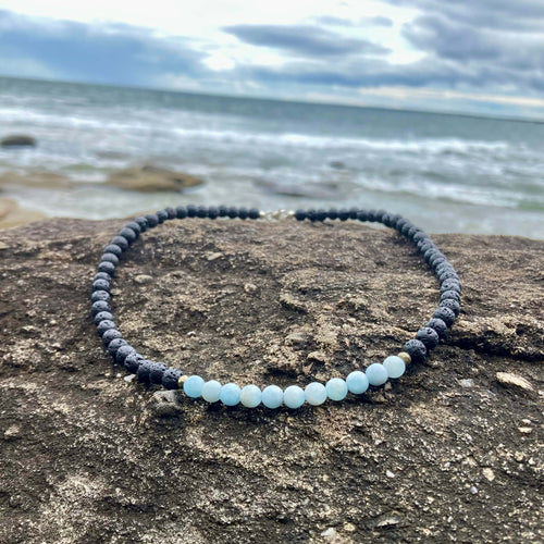 Courage men's necklace: pale blue aquamarine with dark lava stone beads, laid on rock by the sea