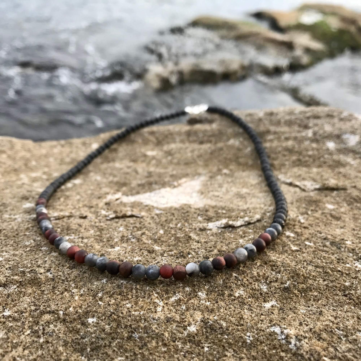 Drive men's necklace: red tiger's eye and grey larvikite beads, on sandstone by the sea