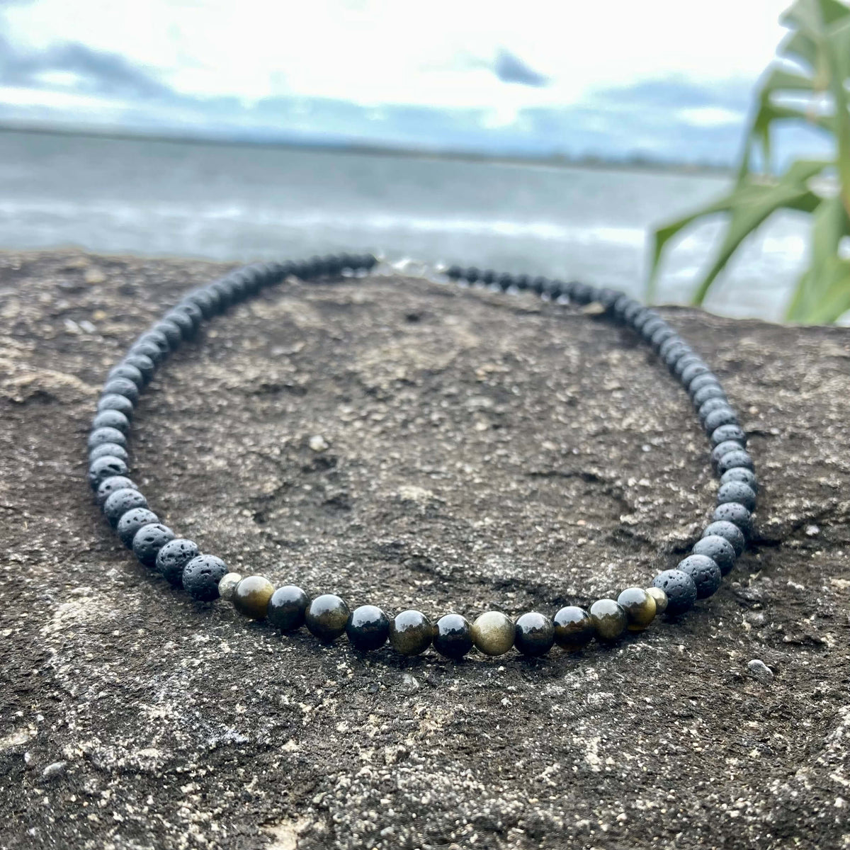 Facing Fears men's necklace: golden obsidian with dark lava stone beads, laid on rock by the sea