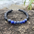 Focus men's bracelet: blue sodalite with matte black lava stone beads, on rock by the sea