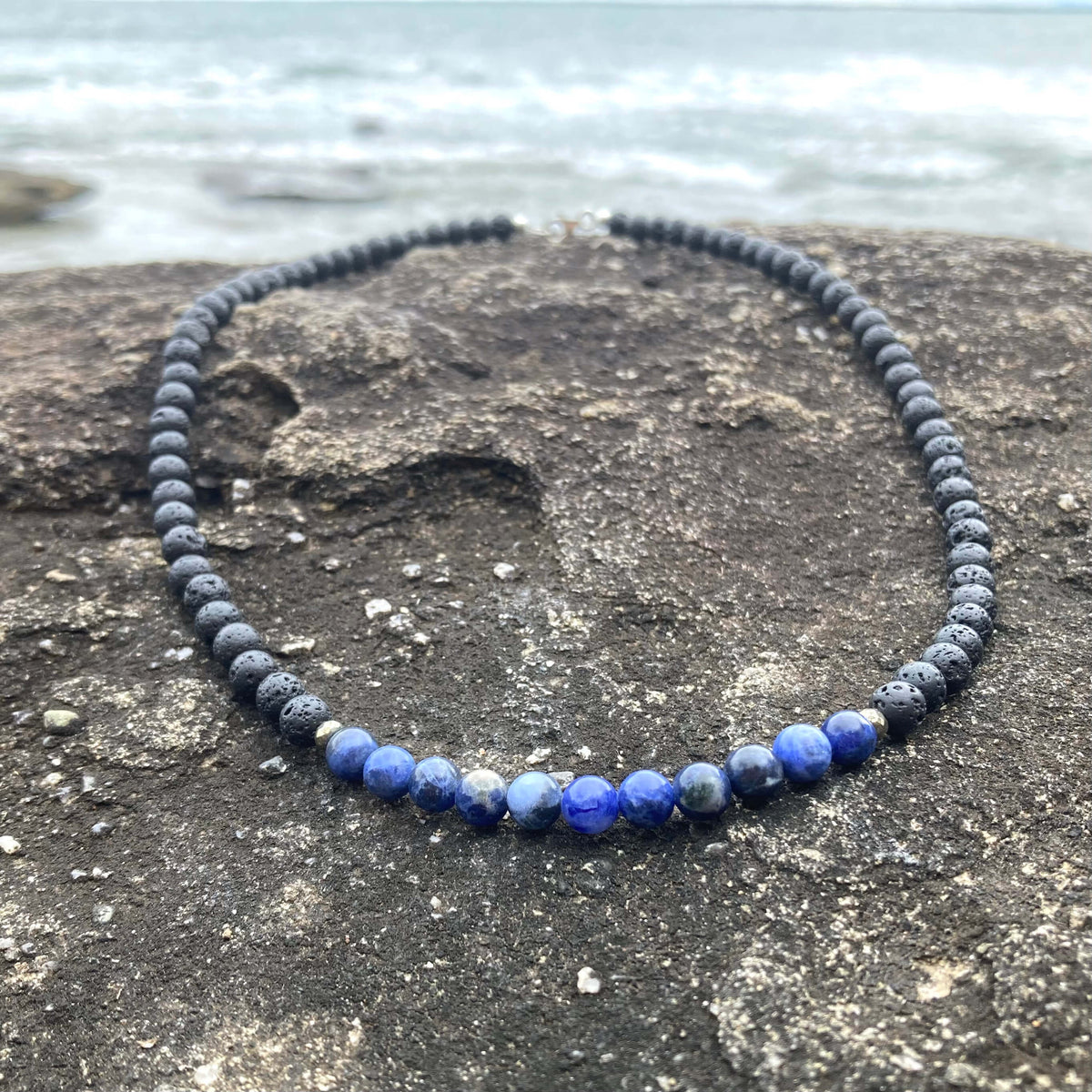 Focus men's necklace: blue sodalite with dark lava stone beads, laid on rock by the sea