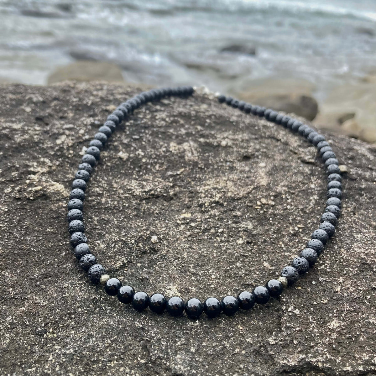 Inner Strength men's necklace: black tourmaline with lava stone and pyrite beads, laid on rock by the sea