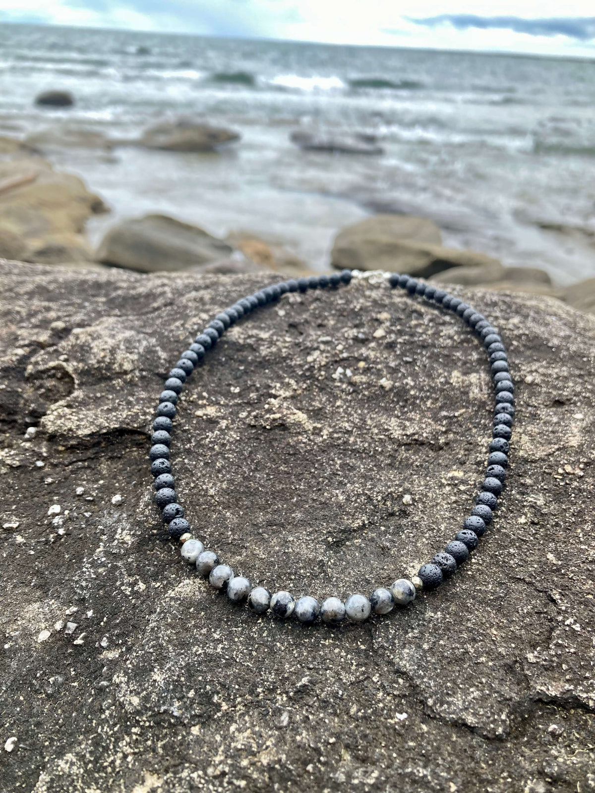 Larvikite section of the Transformation necklace on dark rock with waves behind