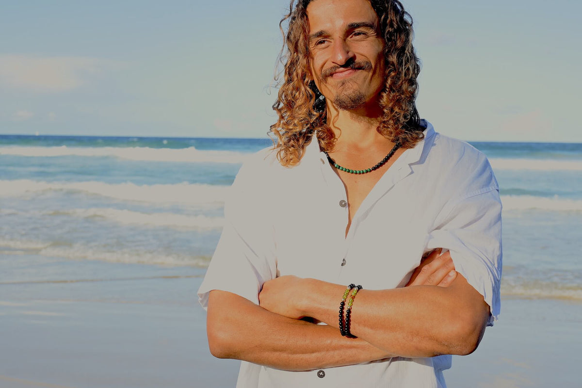 Man standing at the shoreline in golden light, wearing the Manifestation necklace