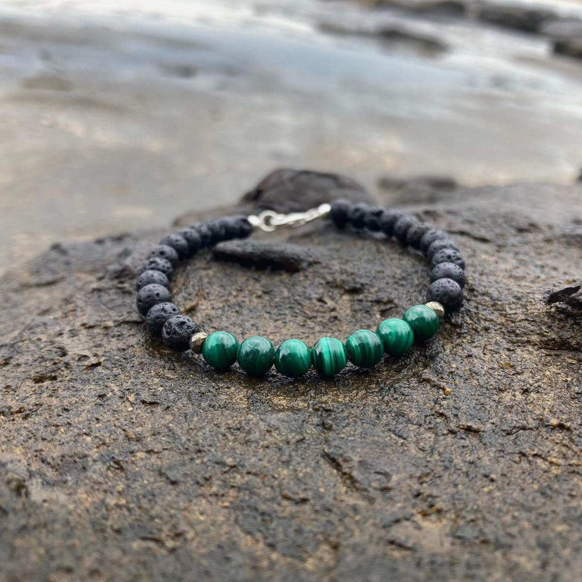 Manifestation men's bracelet: banded green malachite with matte black lava stone beads, on rock by the sea