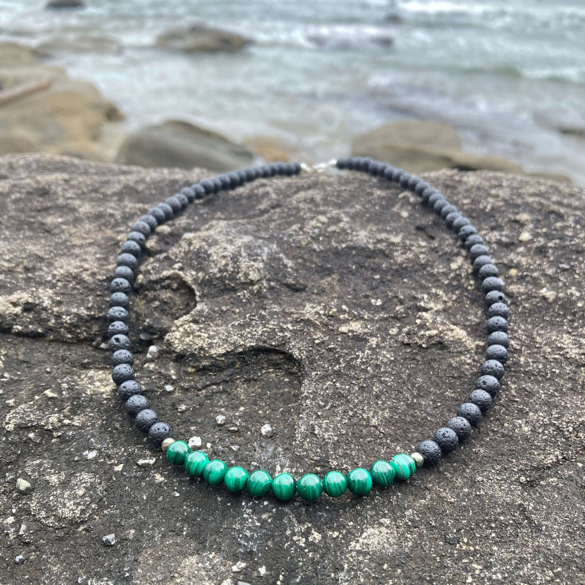 Manifestation men's necklace: green malachite with dark lava stone beads, laid on rock by the sea
