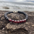 Prosperity men's bracelet: deep red garnet with matte black lava stone beads, on rock by the sea