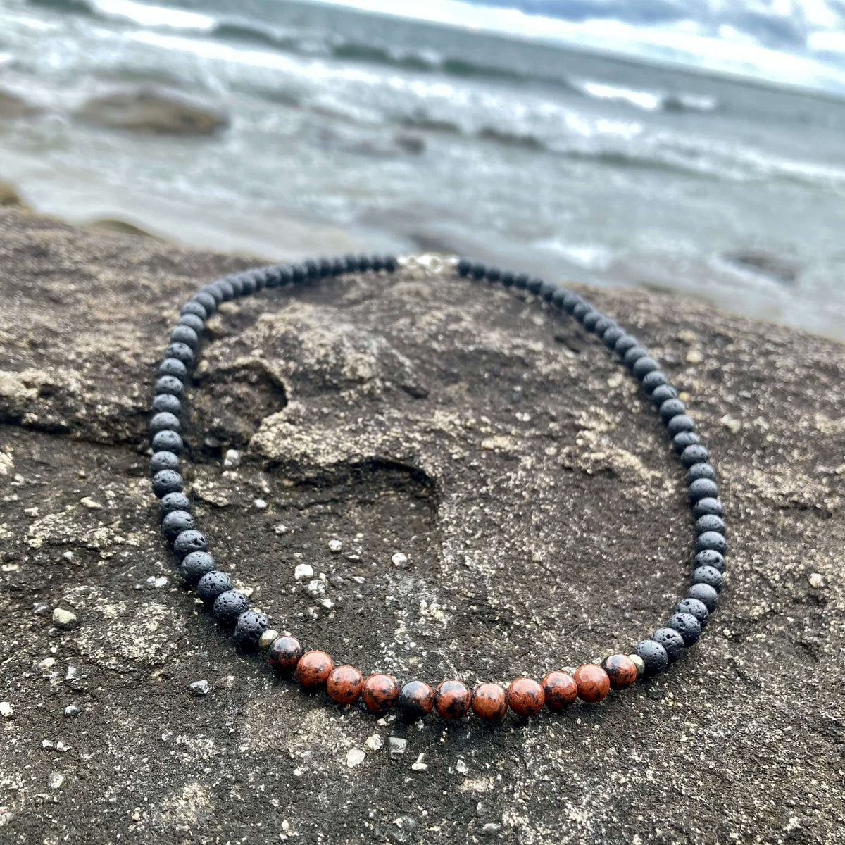 Self-Acceptance men's necklace: red obsidian with dark lava stone beads, laid on rock by the sea