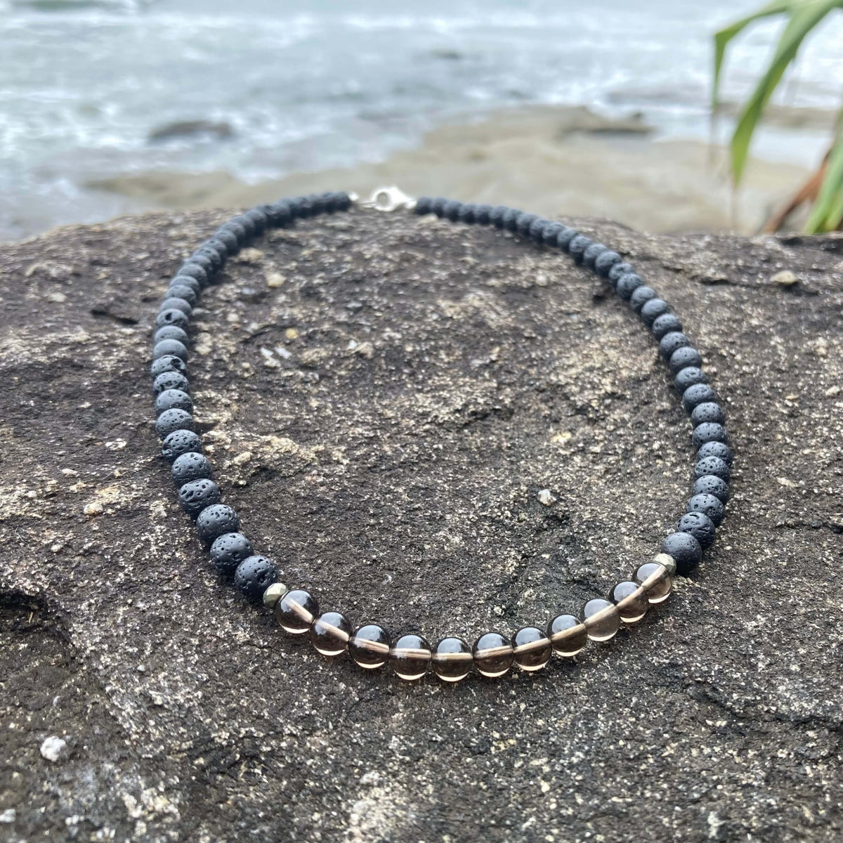Self-Assurance men's necklace: smoky quartz with dark lava stone beads, laid on rock by the sea