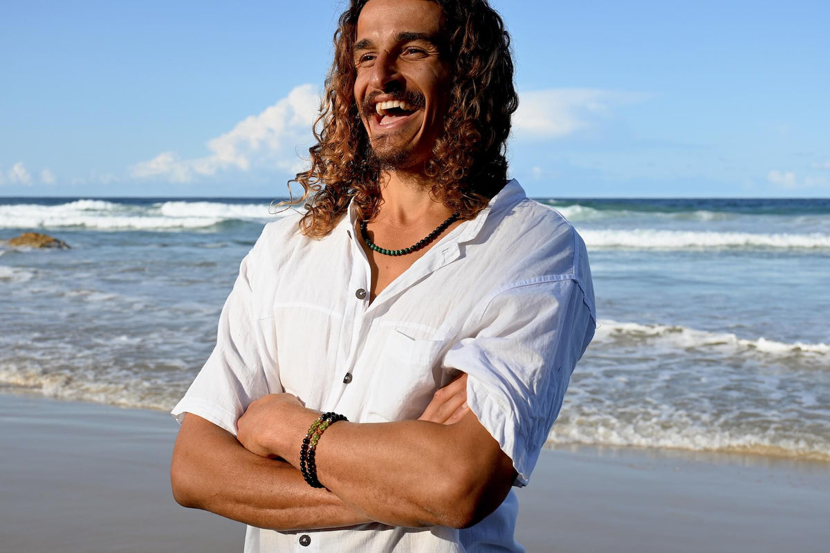 Smiling man in a white shirt at the beach, arms crossed, wearing the Manifestation necklace