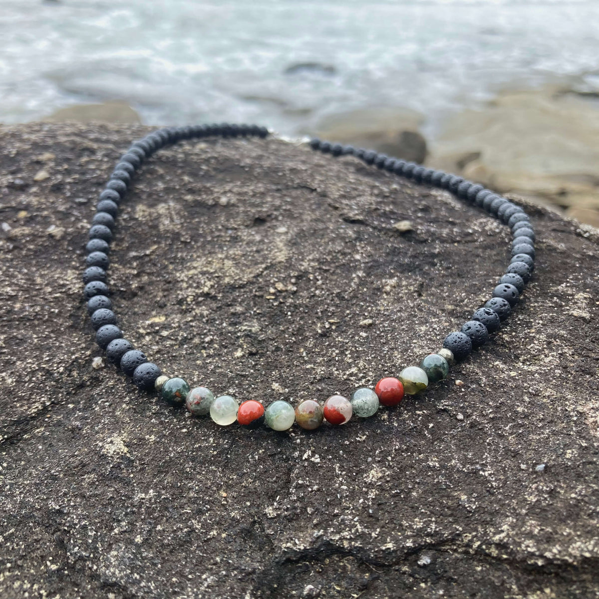 Strength men's necklace: green-and-red bloodstone with dark lava stone beads, laid on rock by the sea