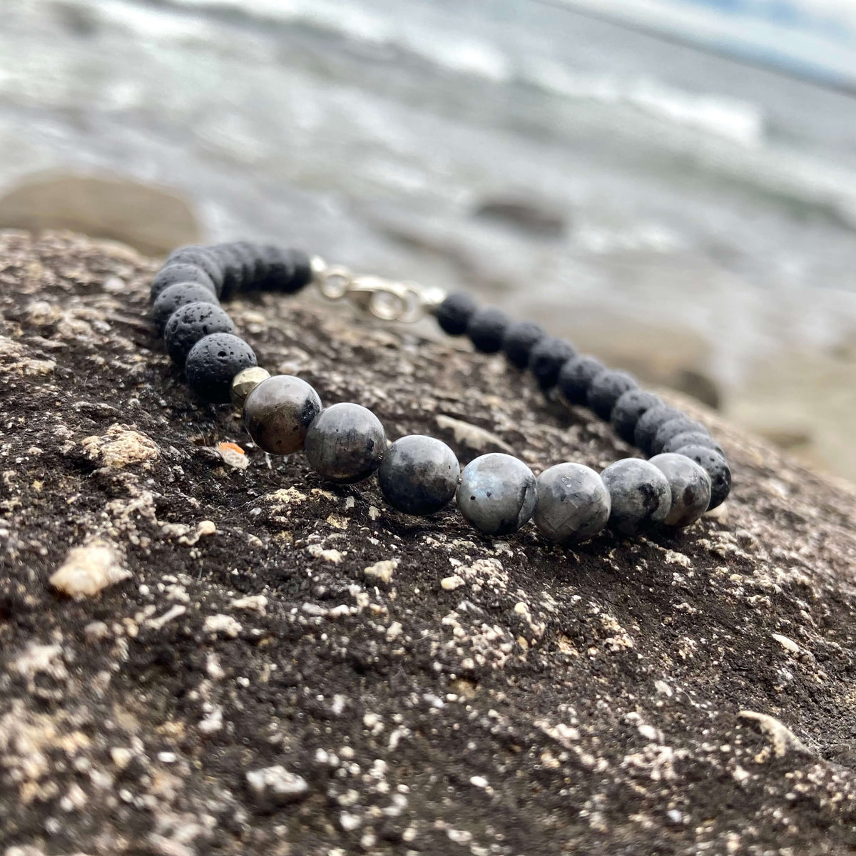 Transformation men's bracelet: grey larvikite with matte black lava stone beads, on rock by the sea