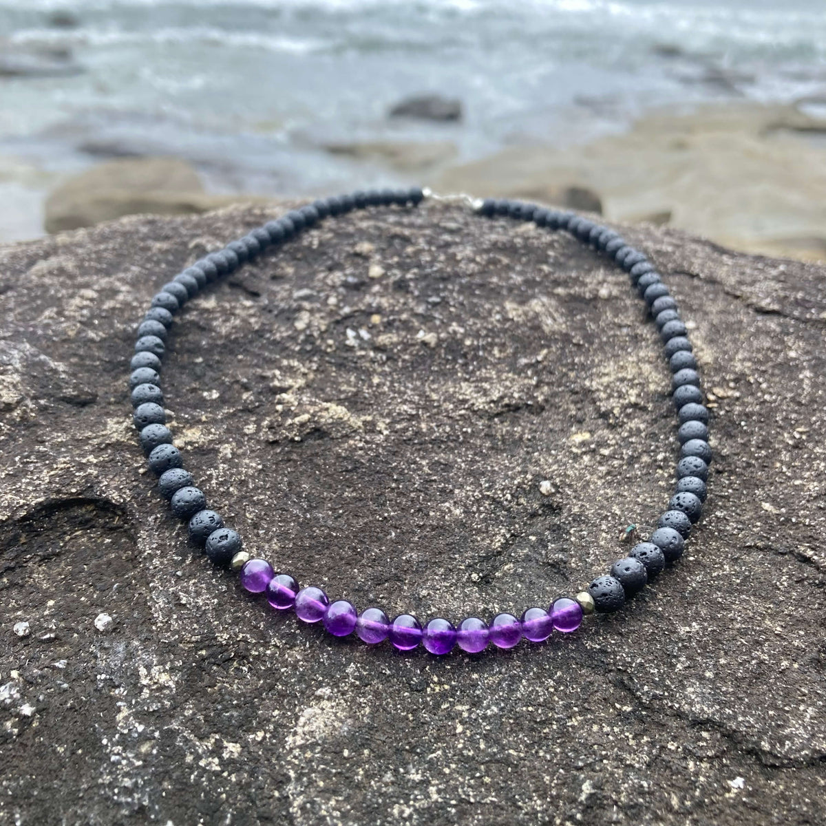 Zen men's necklace: purple amethyst with dark lava stone beads, laid on rock by the sea